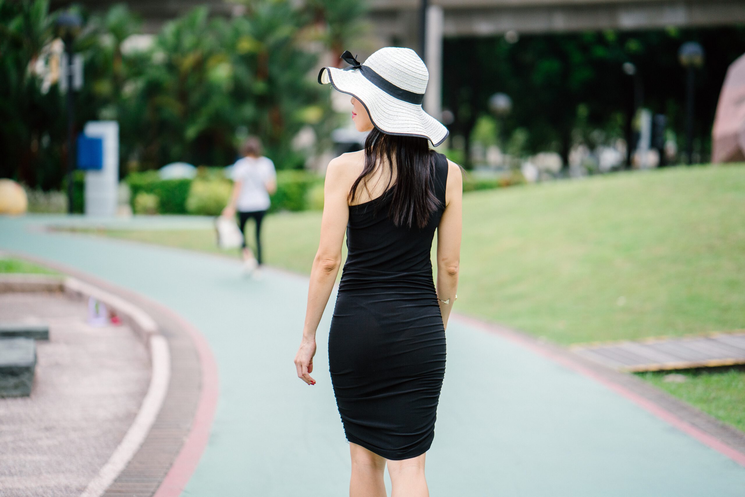 Idąca po chodniku młoda kobieta w czarnej sukience basic i dużym kapeluszu