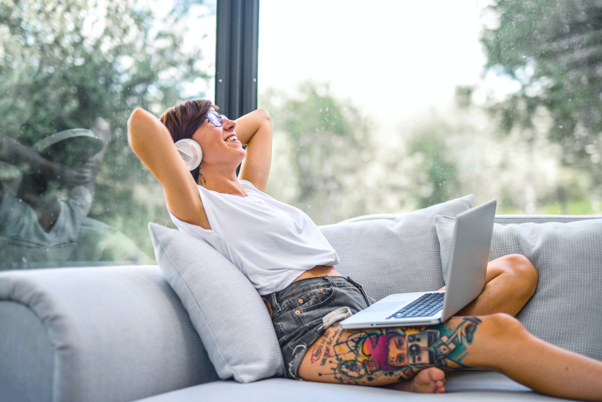 Kobieta w białym topie basic leżąca na sofie z laptopem na kolanach i słuchawkami na uszach