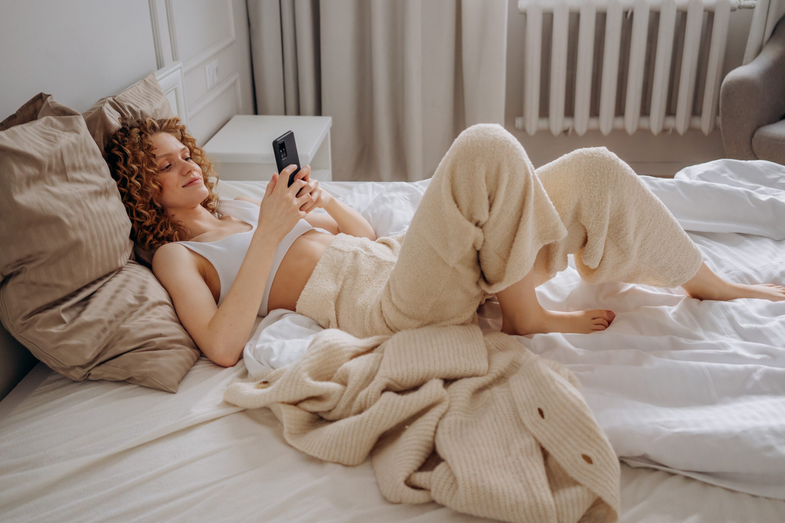 Młoda kobieta w ubraniu basic leżąca na wielkim łóżku i przeglądająca telefon